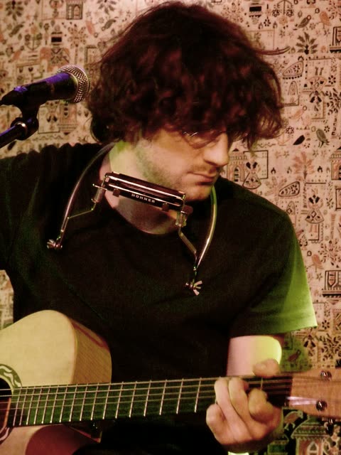 Löhstana David playing harmonica on a neck holder while strumming an acoustic guitar